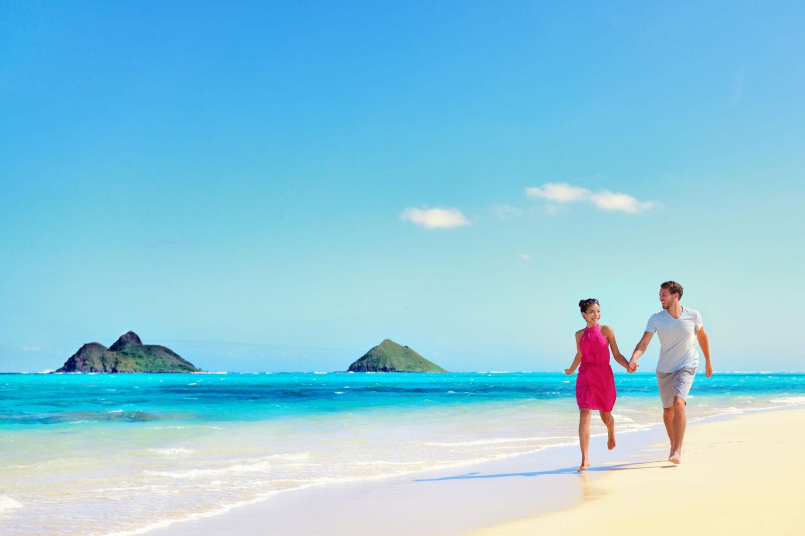 Couple holding hands and strolling on Lanikai Beach with the Mokulua Islands offshore.