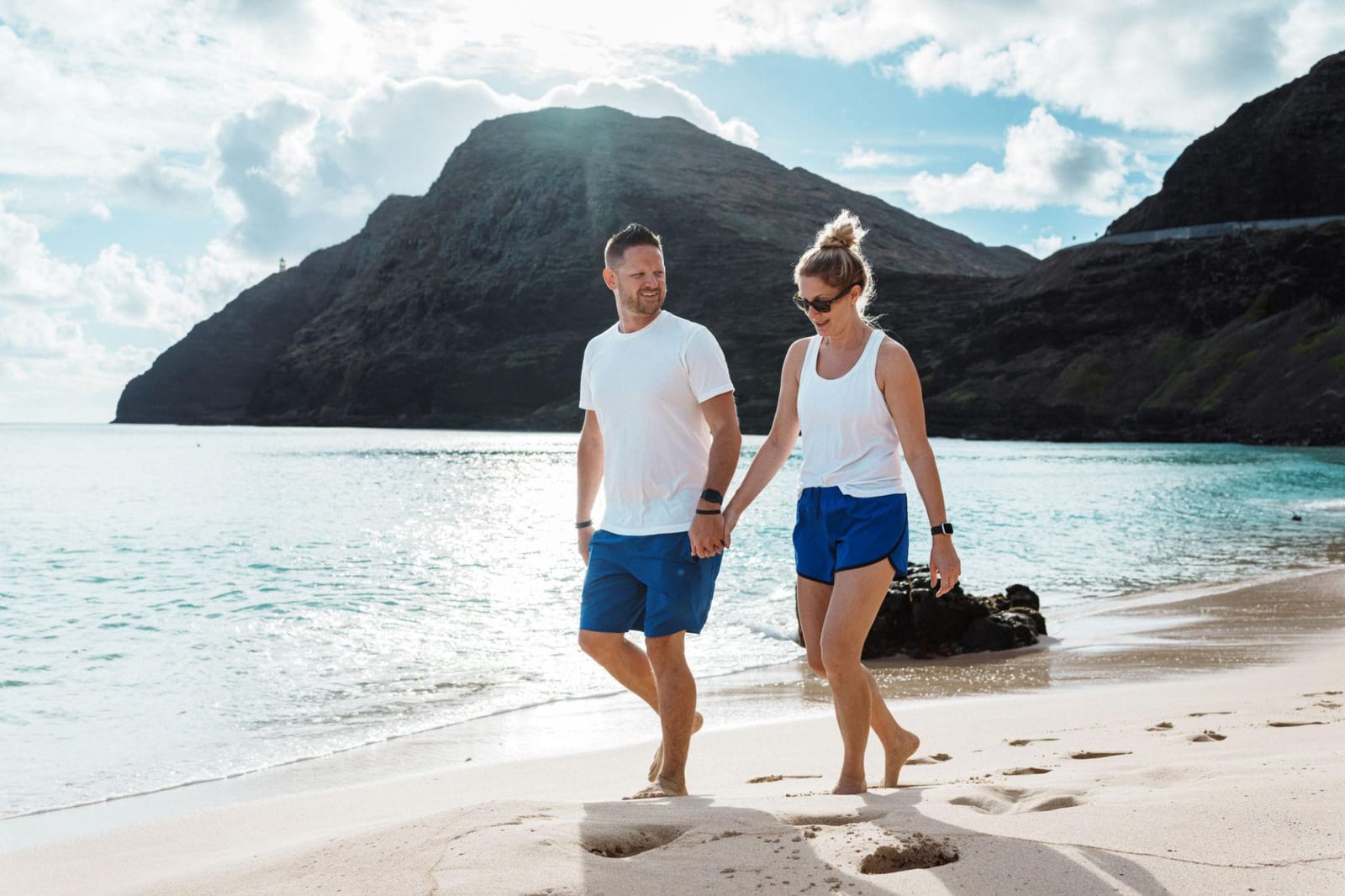 Couple holding hands and walking along the shoreline at Makapu'u Beach on O'ahu, with the lighthouse ridge behind them.