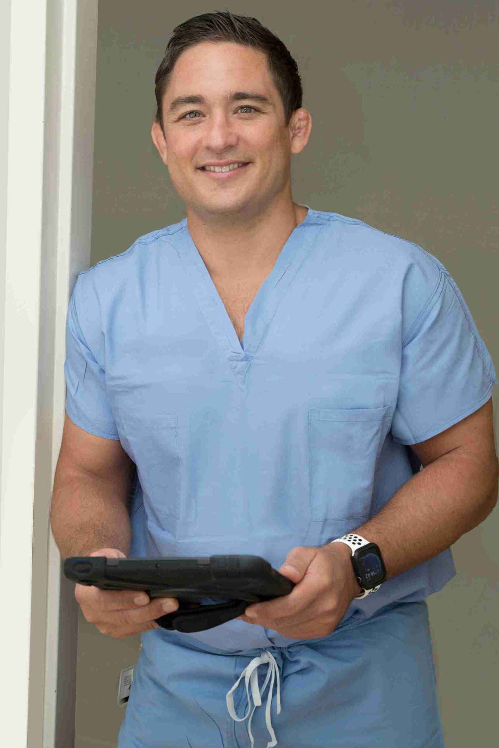 Dr. Paul Morton in blue scrubs smiling while holding a tablet at his Honolulu hip and knee practice.