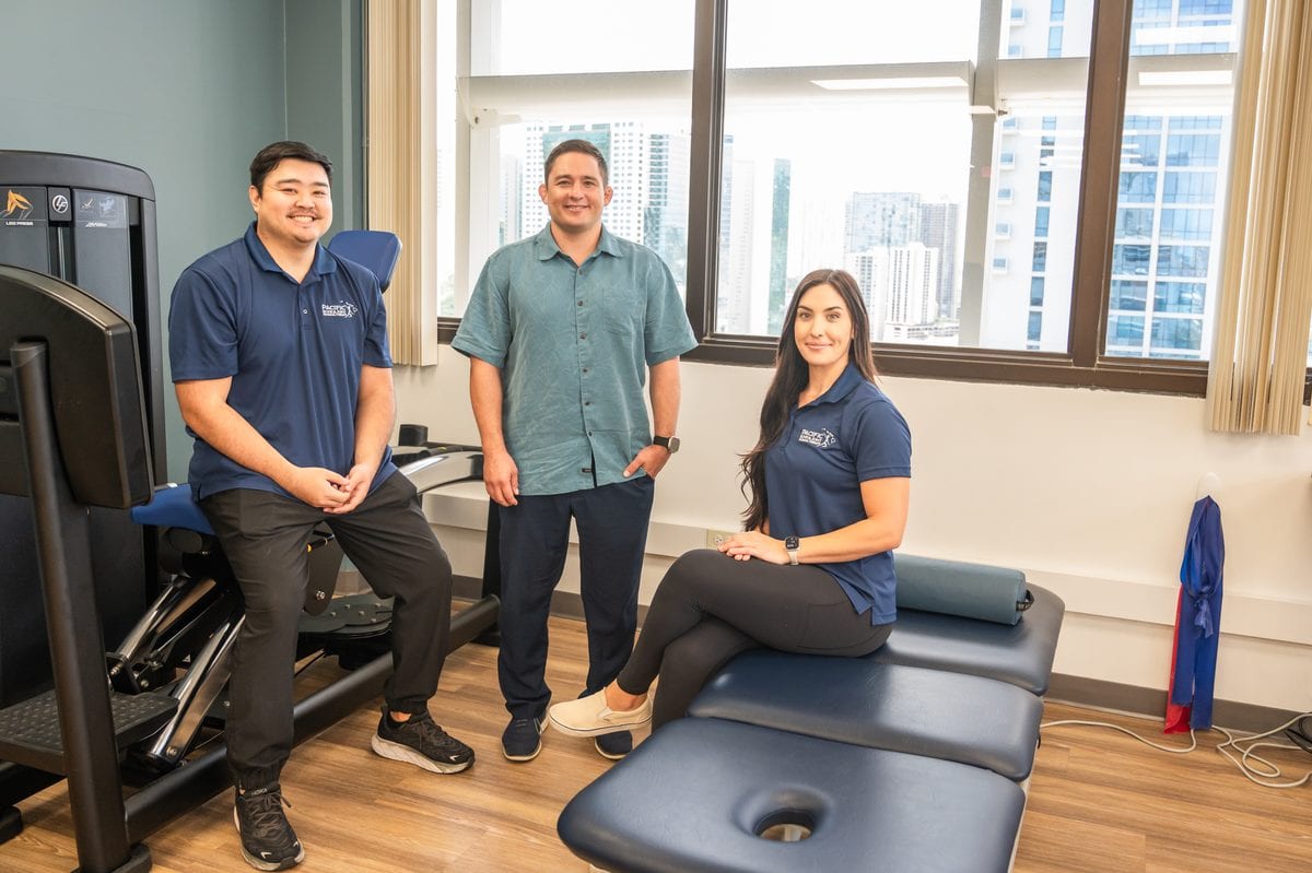 Three PB&J Physical Therapy team members in the Honolulu treatment room with exercise equipment and city views.